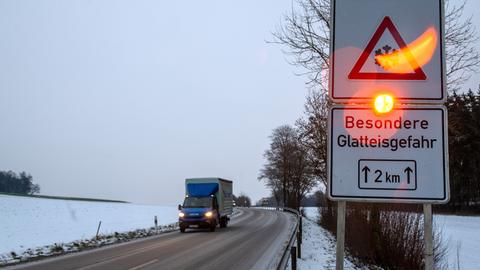 Ein LKW fährt eine verschneite Landstraße entlang. Rechts im Bild steht ein Verkehrsschild mit einer leuchtenden Warnlampe. Das obere Schild zeigt ein Gefahrensymbol mit einer Schneeflocke, und darunter steht: "Besondere Glatteisgefahr" und "2 km". Am Straßenrand sind schneebedeckte Felder und Bäume sichtbar.