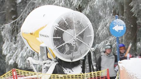 Eine Schneekanone aus der Nähe fotografiert. Im Hintergrund unscharf zwei Skifahrer und Wald mit Schnee in den Bäumen.