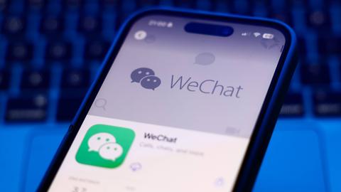 Das Bild zeigt ein Smartphone. In der unteren Hälfte des Bildschirms ist das Symbol der WeChat-App im App-Store zu sehen: ein grünes Viereck, in dem sich zwei Sprechblasen mit Augen befinden. Die obere Hälfte des Bildschirms ist grau eingefärbt, in der Mitte befindet sich der Schritzug "WeChat". Im Hintergrund des Smartphones ist eine Laptop-Tastatur zu sehen.