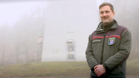 Ein Mann in einer Uniform, auf welcher "Forst" steht. Im Hintergrund der Sockel eines Windrads und Wald.