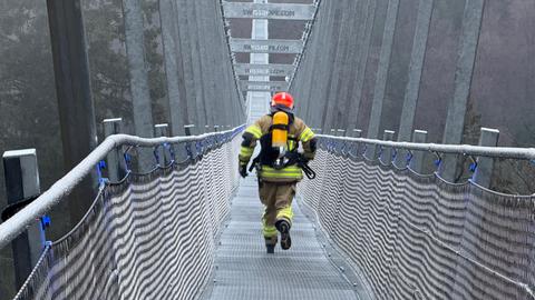 Ein Feuerwehrmann in voller Montur läuft über die Hängebrücke in Willingen. Er ist von hinten zu sehen. Rechts und links sind im Nebel Bäume zu erkennen.