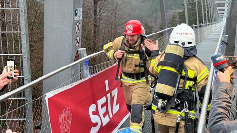 Zwei Feuerwehrleute auf einer hohen Hängebrücke für Fußgänger. Sie sind in voller Montur und klatschen sich ab.