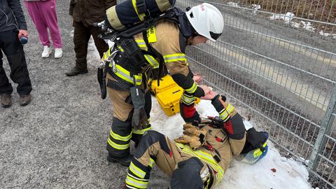 Ein Feuerwehrmann liegt erschöpft auf dem Rücken, eine Kameradin berührt seine Hände. Beide sind in voller Montur.