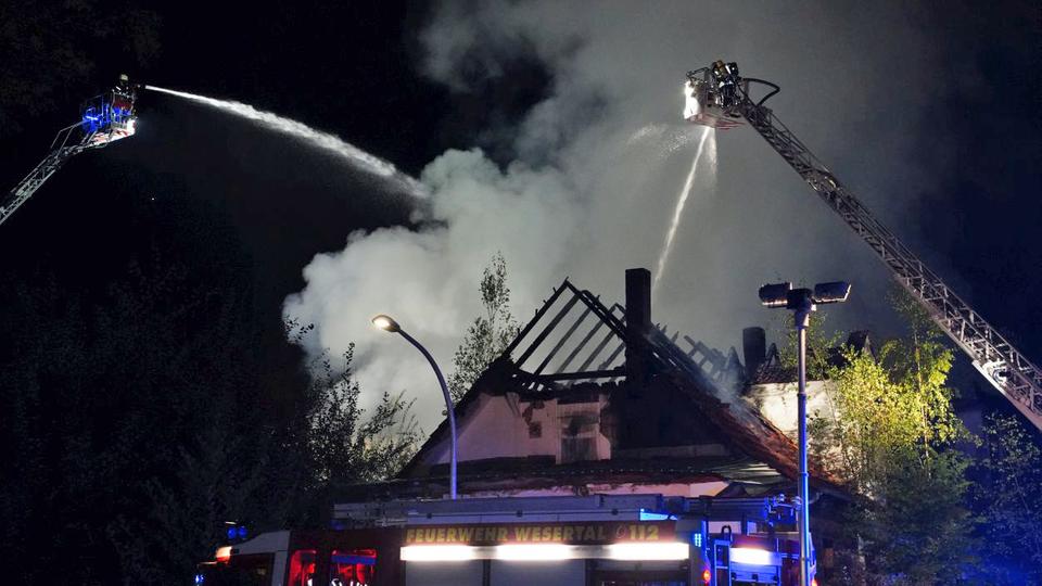 Rauch steigt nachts aus einem Hausdach, bei dem nur noch die Dachbalken stehen. Die Feuerwehr löscht mit Drehleitern.