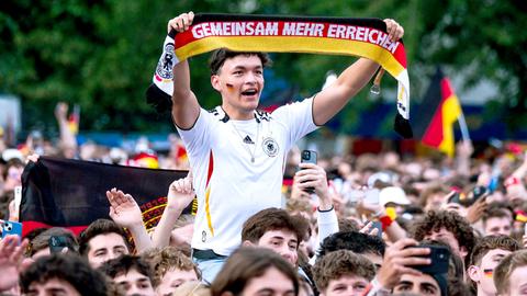 Foto einer Menschenmenge. In der Mitte ein junger Mann erhöht in einem Fußballtrikot, einen Schal in Deutschlandfarben hochhaltend, auf welchem steht: "Gemeinsam mehr erreichen"