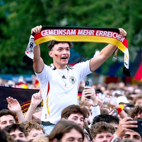 Foto einer Menschenmenge. In der Mitte ein junger Mann erhöht in einem Fußballtrikot, einen Schal in Deutschlandfarben hochhaltend, auf welchem steht: "Gemeinsam mehr erreichen"