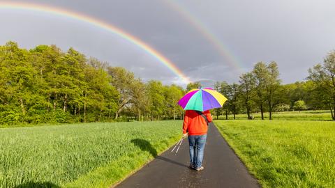 Ein Mann läuft mit einem bunten Regenschirm einen Feldweg entlang. Am Horizont ist ein Regenbogen zu sehen. 