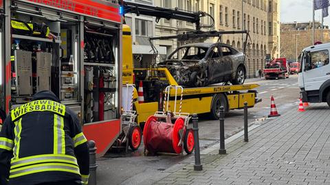 Ein gelbes Abschleppfahrzeug hat auf seiner Ladefläche ein ausgebranntes Wrack eines E-Autos geladen, er steht damit in einer Straße mit Häusern mit Steinfassaden