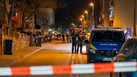  Polizeiwagen und Polizisten in einer aufsteigenden Straße mit Mehrfamilienhäusern hinter einem Polizeiabsperrband bei Nacht