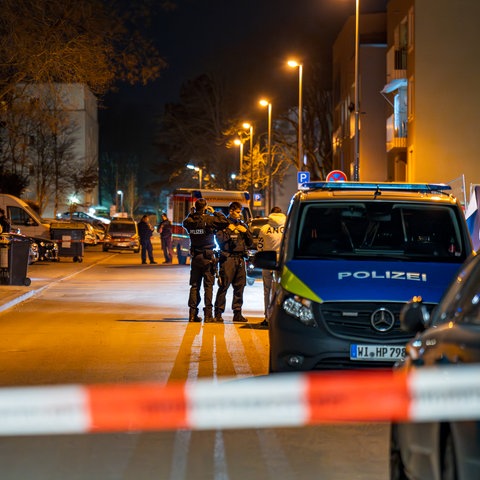  Polizeiwagen und Polizisten in einer aufsteigenden Straße mit Mehrfamilienhäusern hinter einem Polizeiabsperrband bei Nacht