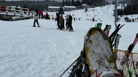 Ein verschneiter Skihang mit mehreren Skifahrern im Hintergrund. Im Vordergrund sind Skier mit Schnee bedeckt. Im Hintergrund sind Bäume, Gebäude und ein Skilift zu sehen.