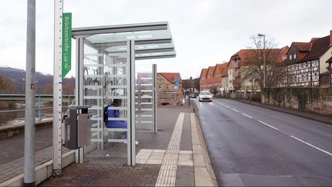 Eine Bushaltestelle ist links im Foto zu sehen,  in der jemand sitzt. Ein Auto fährt auf der Straße daneben. 