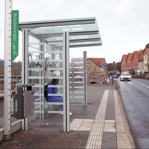 Eine Bushaltestelle ist links im Foto zu sehen,  in der jemand sitzt. Ein Auto fährt auf der Straße daneben. 