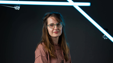 Eine Frau mit langen dunkelblonden Haaren und Brille sitzt vor einer grünen Wand in einem Fotostudio. Sie lächelt in die Kamera und trägt ein zimtrotes Oberteil.