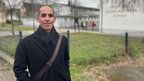 Ein Mann mit Halbglatze und kurzen grauen Haaren an den Kopfseiten steht im schwarzen Mantel vor einem Gebäude., auf dem "Georg-Büchner-Schule" steht. Er lächelt in die Kamera.