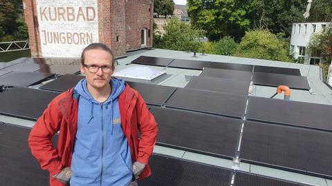 Ein junger Mann sitzt auf einem Dach voller Solarpaneele und schaut ernst in die Kamera