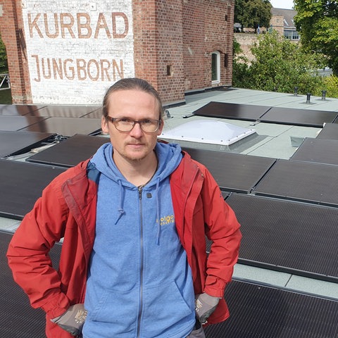 Ein junger Mann sitzt auf einem Dach voller Solarpaneele und schaut ernst in die Kamera