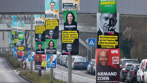 Wahlplakate von verschiedenen Parteien an Laternenmasten auf einer Verkehrsinsel in Frankfurt. Das Bild ist so aufgenommen, dass man die übereinander gehängten Wahlplakate perspektivisch hintereinander in einer Reihe sehen kann.