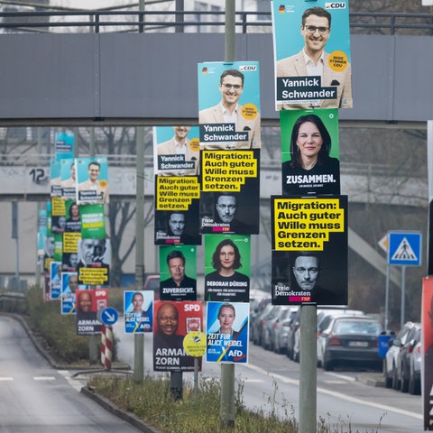 Wahlplakate von verschiedenen Parteien an Laternenmasten auf einer Verkehrsinsel in Frankfurt. Das Bild ist so aufgenommen, dass man die übereinander gehängten Wahlplakate perspektivisch hintereinander in einer Reihe sehen kann.