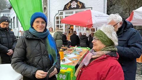 Eine Frau mit Brille und Mütze und Winterjacke lächelt in die Kamera. Sie steht zwischen Leuten in Winterkleidung auf einem Platz vor einer grünen Fahne und einem Biertisch mit Wahlkampfmaterial der Grünen, im Hintergrund ein weißes Haus.