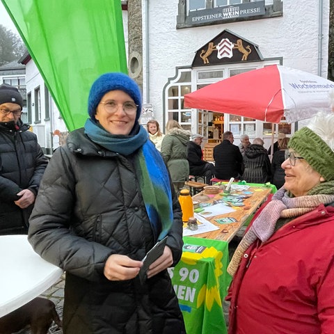 Eine Frau mit Brille und Mütze und Winterjacke lächelt in die Kamera. Sie steht zwischen Leuten in Winterkleidung auf einem Platz vor einer grünen Fahne und einem Biertisch mit Wahlkampfmaterial der Grünen, im Hintergrund ein weißes Haus.