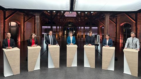 Vier Frauen und drei Männer stehen hinter Stehpulten in einem Halbkreis in einem großen Veranstaltungssaal mit Backsteinmauern. Sie schauen in die Kamera.