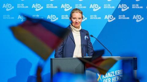 Die AfD-Vorsitzende Alice Weidel steht mit zurückgebundenen blonden Haaren und im dunklen Blazer hinter einem Rednerpult auf einer Wahlkampfbühne in einer Halle. Hinter ihr eine Wand mit hellblauer Tapete und vielen "AfD"-Logos.