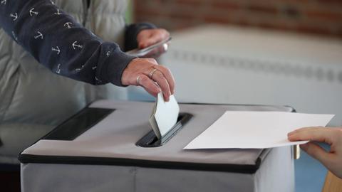 Eine Hand steckt einen Zettel in eine graue Wahlurne. Im Hintergrund unscharf ein Wahllokal.