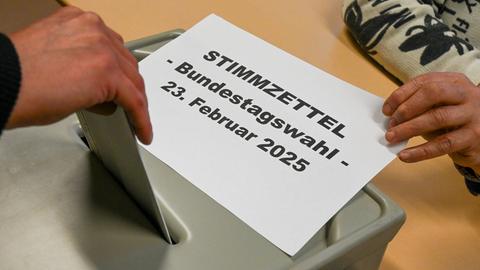 Eine Hand steckt einen Zettel in eine graue Wahlurne. Im Hintergrund unscharf ein Wahllokal.