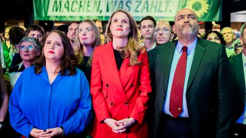 Das Bild zeigt Die Grünen-Vorsitzenden Ricarda Lang (links) und Omid Nouripour (rechts) mit Spitzenkandidatin Terry Reintke bei einer Wahlparty der Grünen zur Europawahl. Hinter ihnen sind Parteimitglieder sowie ein Banner mit der Aufschrift "Machen, was zählt" zu sehen. Alle Blicken mit ernstem Gesicht auf einen Bildschirm.