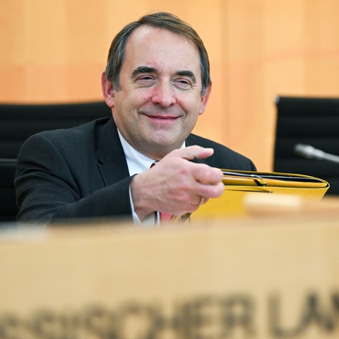 Ein Mann sitzt an einem Pult mit der Aufschrift "Hessischer Landtag" und hält lächelnd einen Ordner in der Hand.