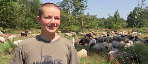 Eine junge Frau lächelt in die Kamera. Hinter ihr ist eine Schafherde auf einer Wiese zu sehen.