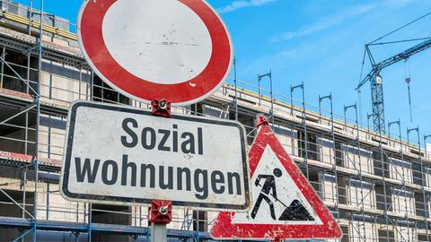 Schilder an einer imaginaeren Baustelle. Verkehrszeichen 250 (Verbot fuer Fahrzeuge aller Art) in runder Form, ein Zusatzschild mit der Aufschrift: "Sozialwohnungen" an einem Schildermast und im Hintergrund das Schild Zeichen 123 Arbeitsstelle bzw. Baustelle vor einem Mehrfamilenhaus-Rohbau mit Baugeruesten. 