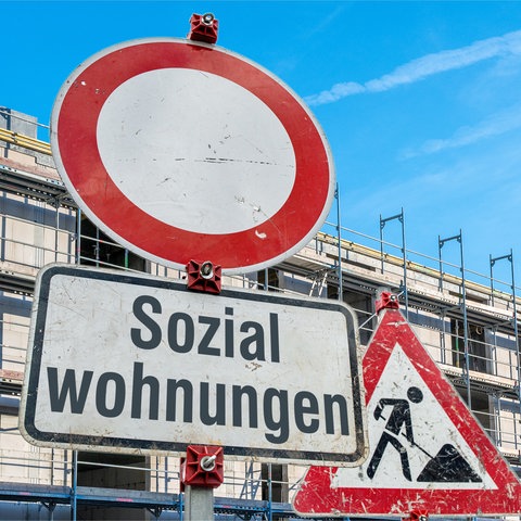 Schilder an einer imaginaeren Baustelle. Verkehrszeichen 250 (Verbot fuer Fahrzeuge aller Art) in runder Form, ein Zusatzschild mit der Aufschrift: "Sozialwohnungen" an einem Schildermast und im Hintergrund das Schild Zeichen 123 Arbeitsstelle bzw. Baustelle vor einem Mehrfamilenhaus-Rohbau mit Baugeruesten. 