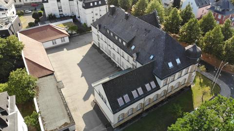 Zu sehen ist eine Luftaufnahme des Rathauses Friedberg. Wir schauen herunter auf ein weiß-braunes, herrschaftliches Gebäude mit einem schwarzen Dach. Davor ein leerer Betonparkplatz, drumherum Wohnhäuser und grüne Bäume