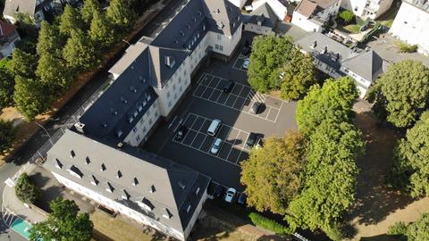 Zu sehen ist eine Luftaufnahme des Rathauses Friedberg. Wir schauen herunter auf ein weiß-braunes, herrschaftliches Gebäude mit einem schwarzen Dach. Davor ein leerer Betonparkplatz, drumherum Wohnhäuser und grüne Bäume