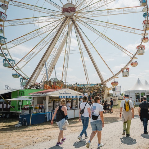 Das Bild zeigt Menschen, die über das Hessentagsgelände gehen. Im Hintergrund ist ein Riesenrad und Verkaufsstände zu sehen. 