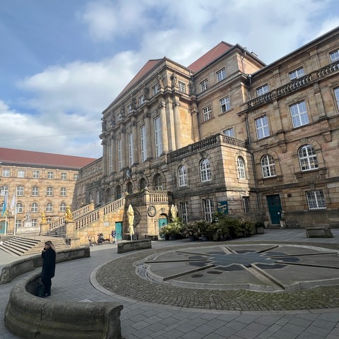 Das Rathaus in Kassel, zu sehen ist die große Treppe aus Stein, rechts und links zwei goldene Löwen. Im Vordergrund ist der Aschrottbrunnen zu sehen.