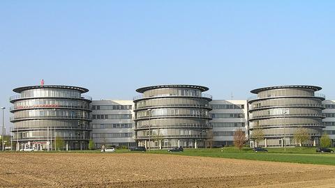 Graues Bürogebäude mit drei kreisförmigen Gebäudeteilen vor einem Gebäuderiegel, davor ein Acker