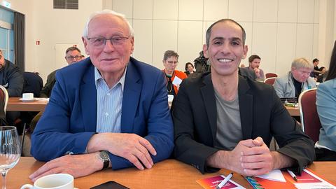 Oskar Lafontaine (l.) und Ali Al-Dailami an einem Tisch sitzend und in die Kamera lächelnd, dahinter andere BSW-Delegierte an Tischen