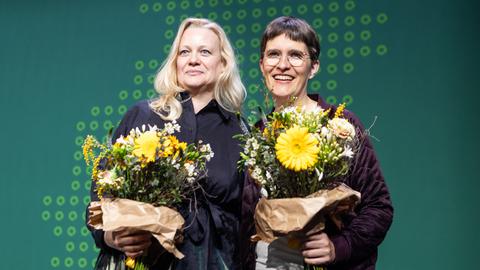 Zwei Frauen mit Blumensträußen in der Hand vor dunkelgrünem Hintergrund, lachen in die Kamera