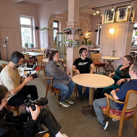 In einem Café in Marburg sitzen vier Menschen um einen Tisch herum. Davor hocken ein Kameramann und eine Kamerafrau und filmen die Gespräche.