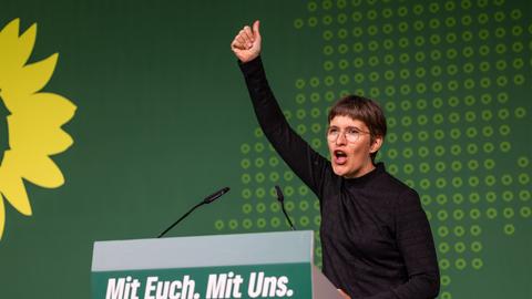 Anna Lührmann am Rednerpult mit der Aufschrift "Mit Euch. Mit Uns. Für Hessen." reckt den rechten Arm hoch mit Daumen-hoch-Zeichen, dahinter grüne Wand mit Sonnenblumen-Logo der Grünen