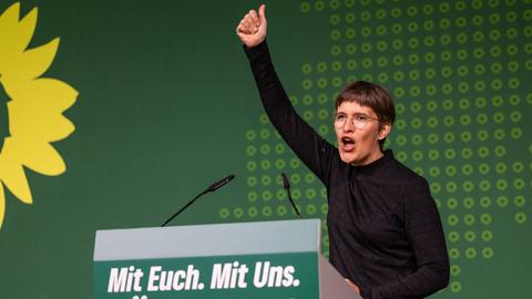 Anna Lührmann am Rednerpult mit der Aufschrift "Mit Euch. Mit Uns. Für Hessen." reckt den rechten Arm hoch mit Daumen-hoch-Zeichen, dahinter grüne Wand mit Sonnenblumen-Logo der Grünen