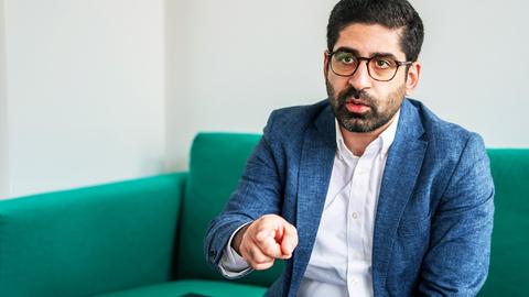 Ein im Anzug spricht und zeigt mit dem Finger. Foto seines Oberkörpers, während er sitzt.