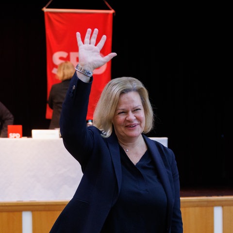 Nancy Faeser (SPD) winkt strahlend den Delegierten einer SPD-Versammlung zu, im Hintergrund drei Delegierte und eine SPD-Fahne