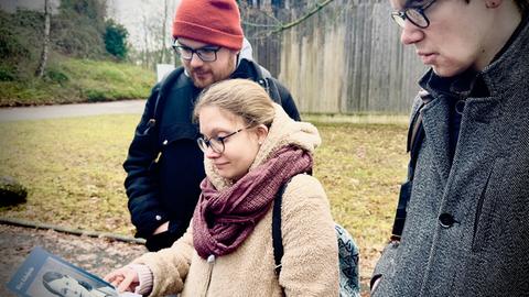 Die Inklusiv-Reporter der Normalos schauen sich das Foto einer Frau an, die in der NS-Tötungsanstalt in Hadamar ermordet wurde. Im Hintergrund ist eine hohe Mauer zu sehen.