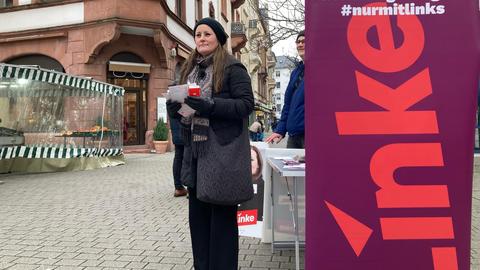 Eine Frau im grauen Mantel und mit einer schwarzen Mütze steht an einem Wahlkampfstand. Auf einem lilafarbenen Aufsteller steht "Die Linke".