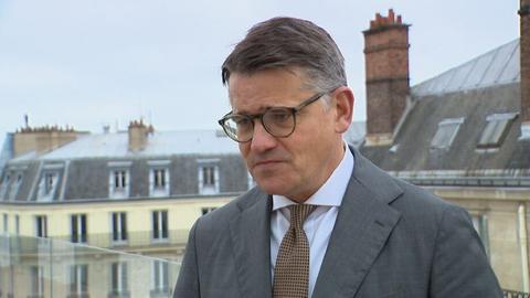 Das Bild zeigt den hessischen Ministerpräsidenten Boris Rhein bei einem Besuch in Paris. Er steht auf einem Balkon, der von einer Glasbalustrade eingegrenzt ist und trägt ein graues Sakko, eine Krawatte mit brauenn Punkten und eine dunkel gerahmte Brille.