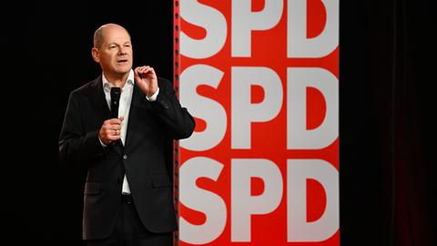 Scholz steht mit Mikro auf Bühne und spricht vor einer SPD-Werbewand.